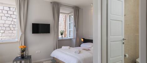 A modern bedroom with a bed, a TV mounted on the wall, a small table with a bottle and glasses, and a window with curtains.