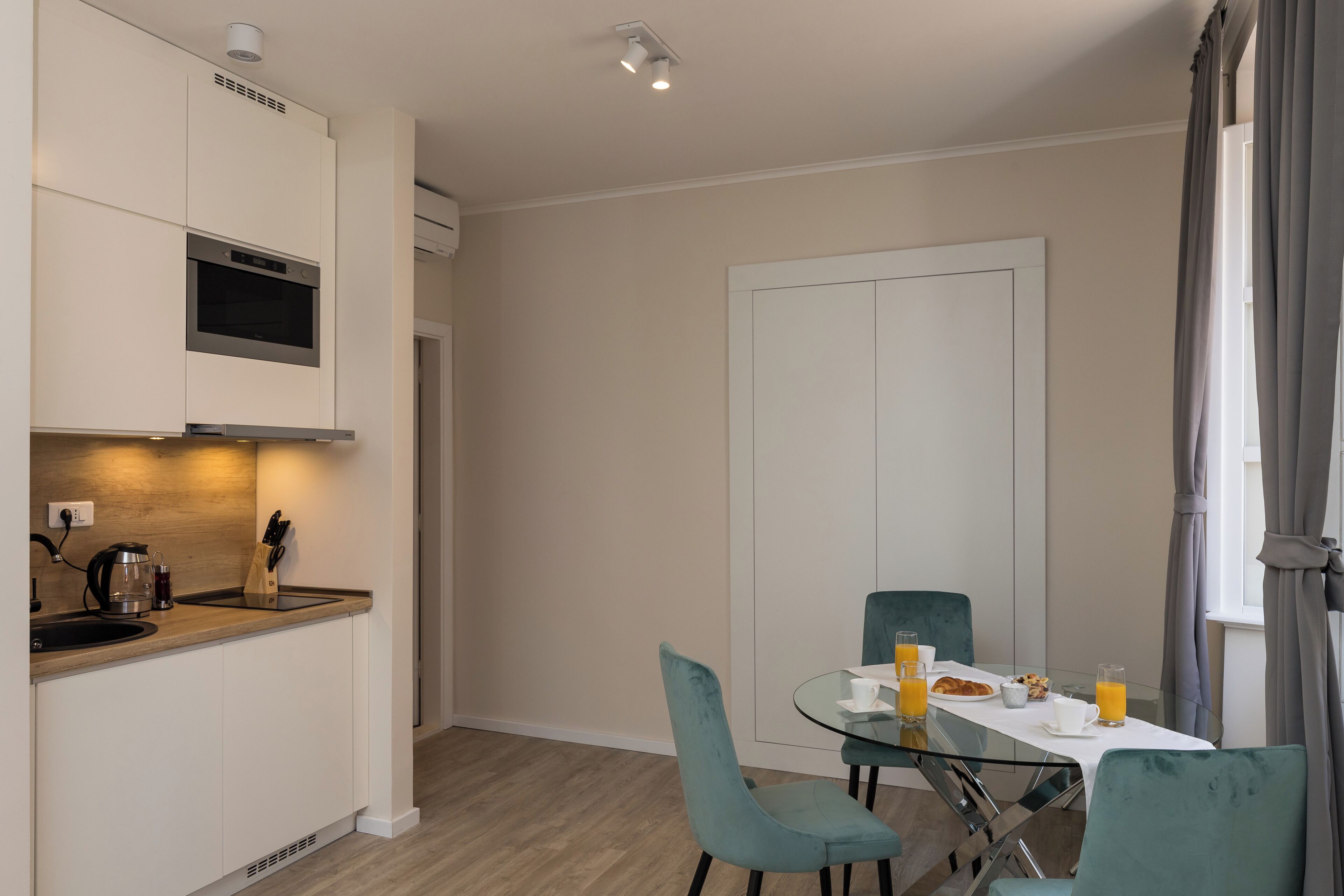 A modern kitchen with built-in appliances, a dining area with a glass table, and teal chairs.