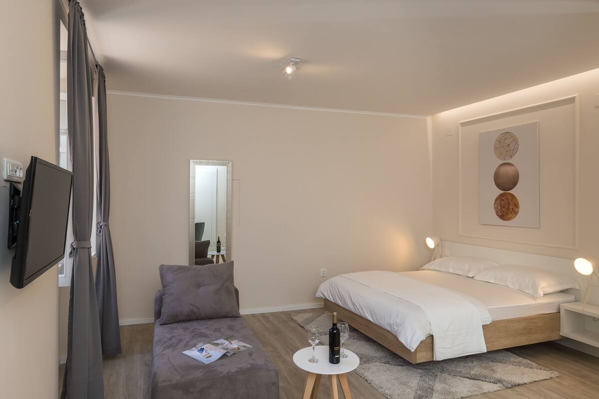 Ein modernes Schlafzimmer mit einem Bett, einem Fernseher, einem kleinen Tisch mit einer Flasche und Gläsern sowie einem grauen Sofa.