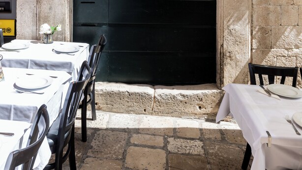 Un espace de restauration en plein air avec des tables prévues pour deux personnes, une porte noire et des murs en pierre.
