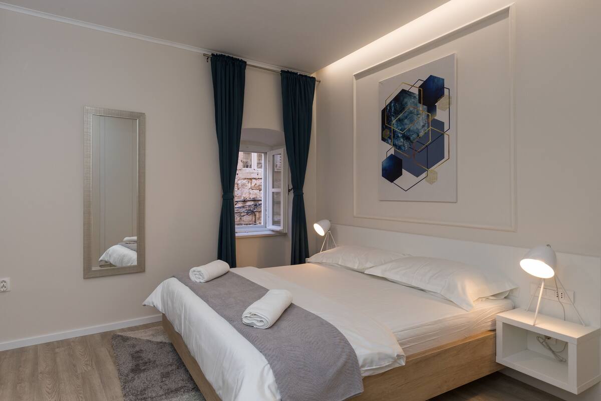 Ein modernes Schlafzimmer mit einem großen Bett, einem Nachttisch mit Lampe, einem Spiegel und einem geometrischen Wandbild.