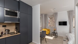 Une cuisine moderne avec des meubles sombres et un four encastré, un coin repas avec une chaise jaune, une petite table d’appoint, et un lit avec une tête de lit blanche dans la pièce adjacente.