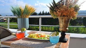 A table with a fruit platter, two drinks, and two vases on a balcony overlooking a scenic view. - Marinn Deluxe Hotel (Altinova)