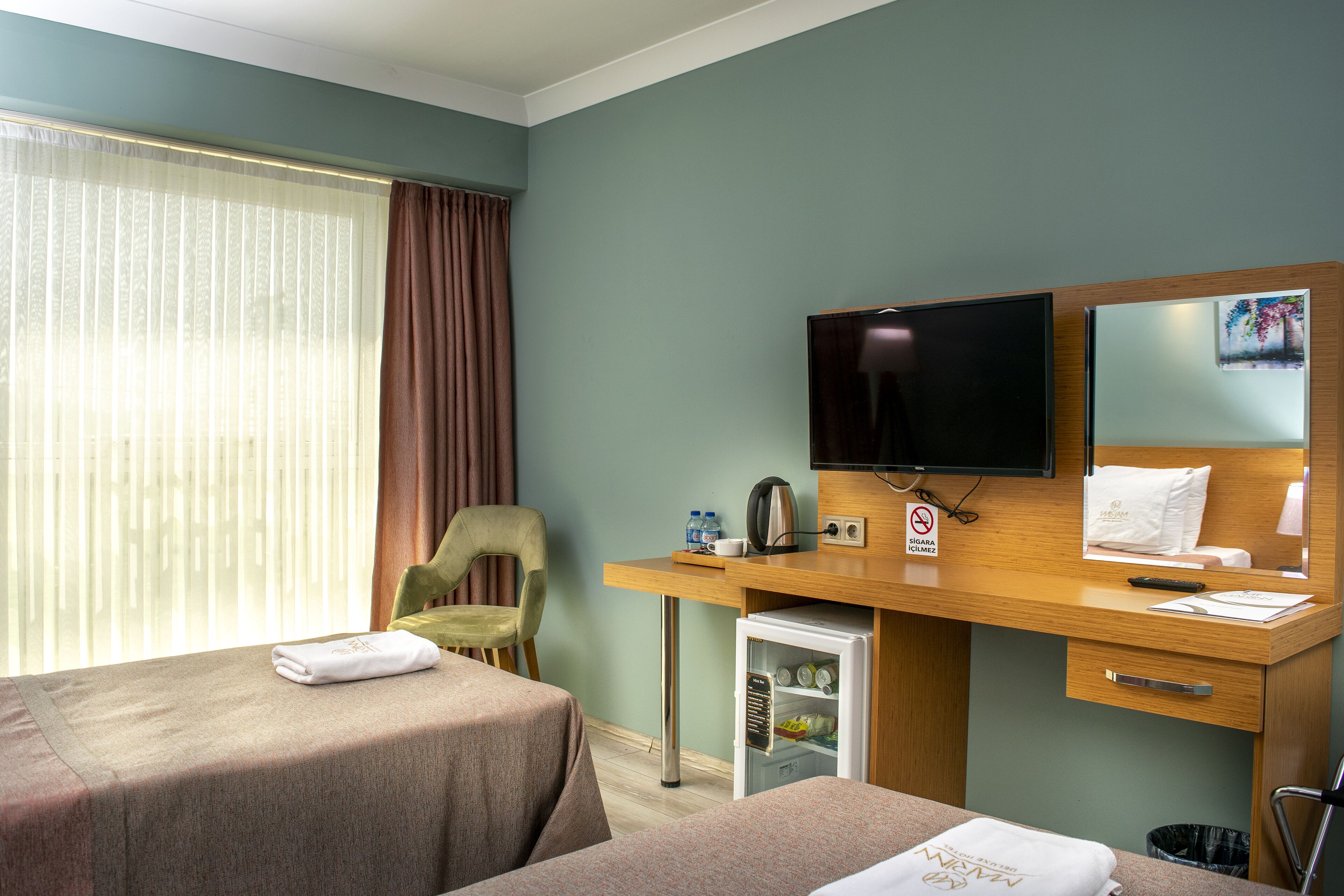 Ein Hotelzimmer mit zwei Betten, einem Schreibtisch, einem Fernseher, einer Minibar und einem Fenster mit Vorhängen.