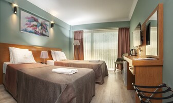 Habitación de hotel con dos camas, un escritorio, una silla, una lámpara, un cuadro y un ventanal grande con cortinas.