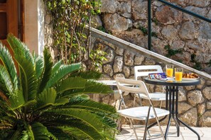 Un área de estar al aire libre con un muro de piedra, sillas blancas, una mesa y un vaso de jugo de naranja.