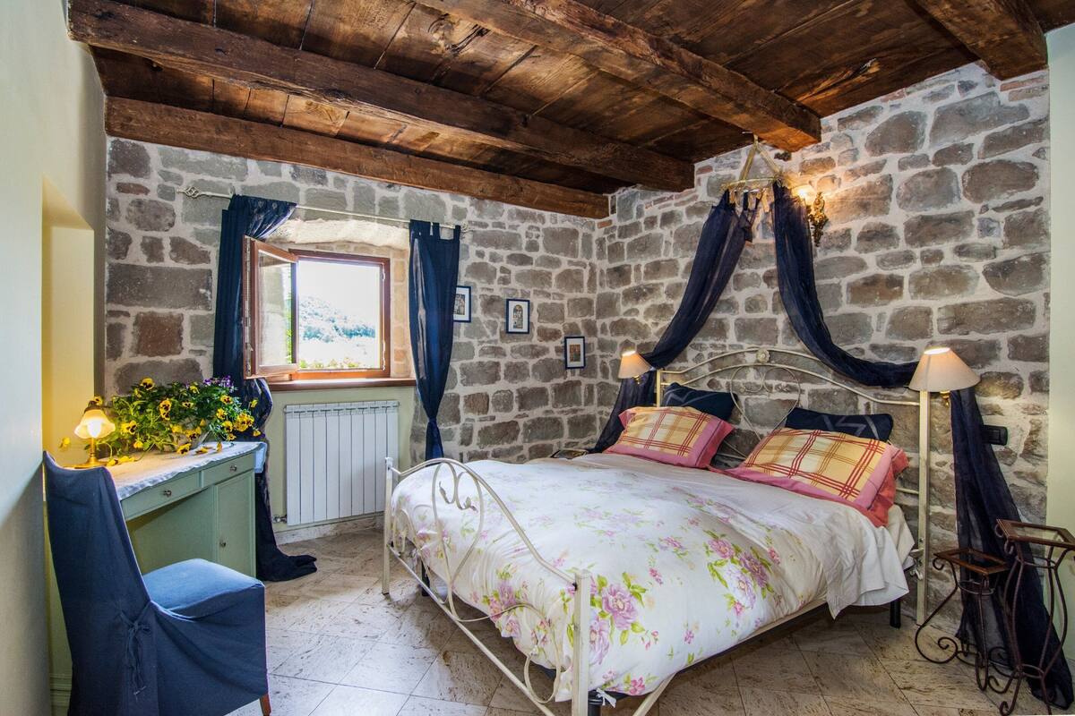 Un dormitorio rústico con pared de piedra, techo de madera, cama con ropa de cama floral, escritorio con lámpara y una silla.