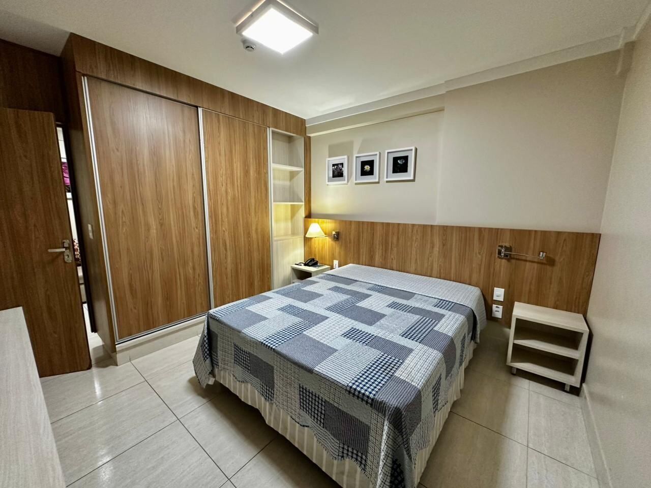 Un dormitorio con cama, armario de madera, mesita de noche, lámpara y una pequeña estantería.