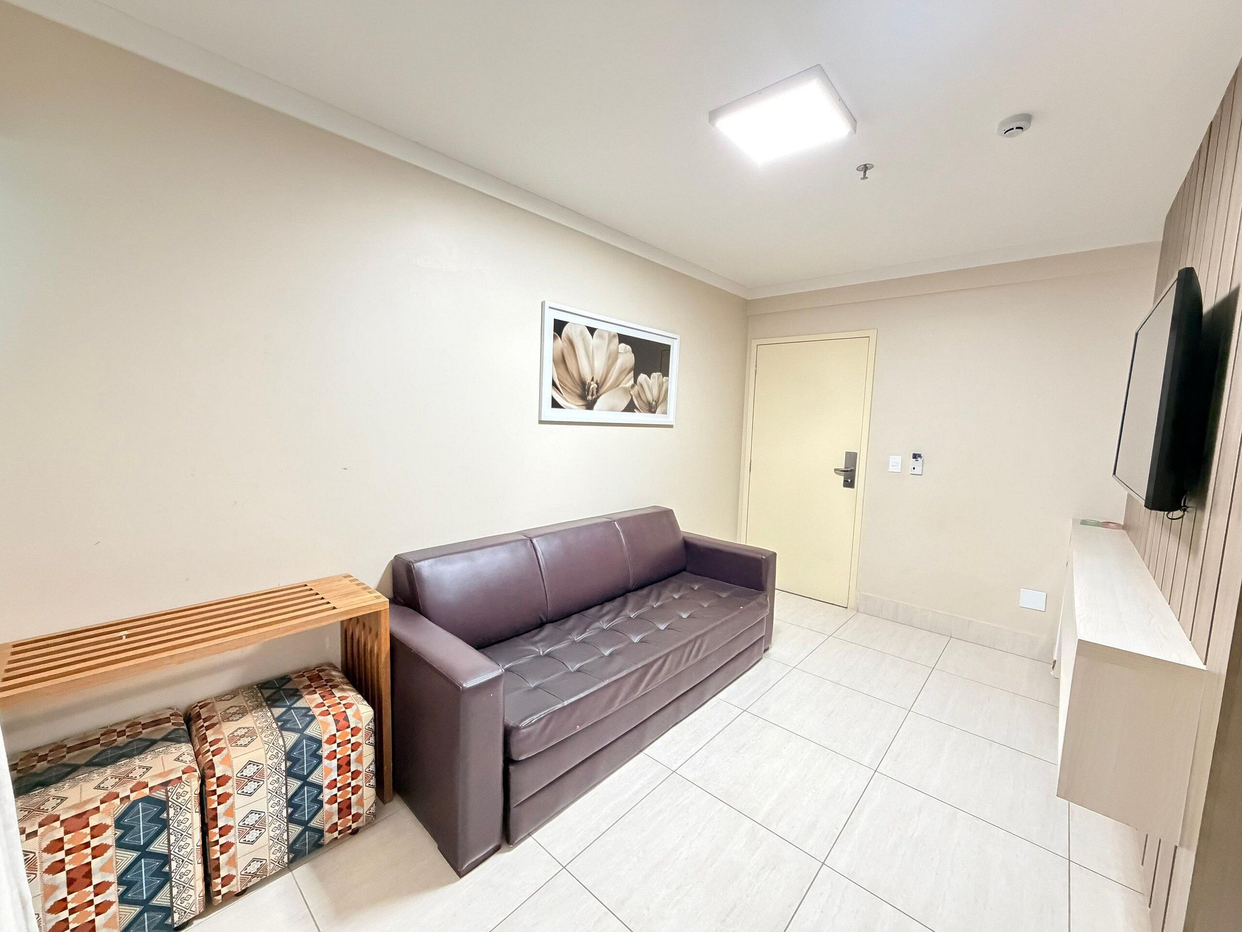 A modern living room with a brown leather sofa, a wooden side table, patterned cushions, a framed picture on the wall, and a door in the background.