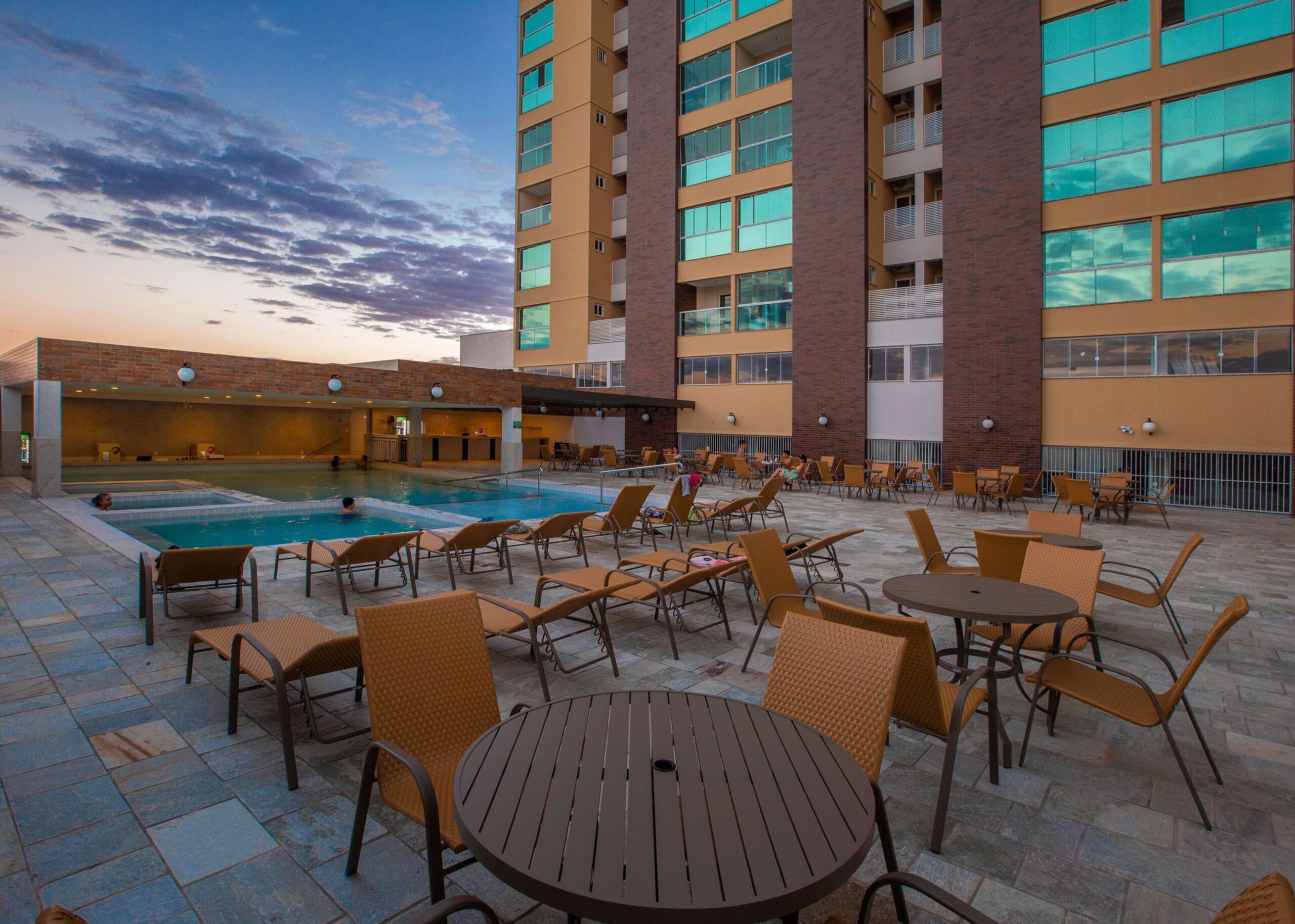A hotel pool area with lounge chairs and tables, a swimming pool, and a building with multiple floors and balconies.