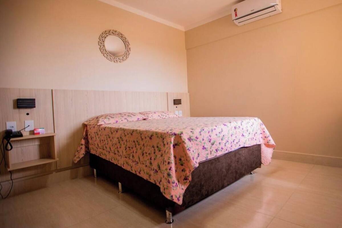 A single bed with a floral bedspread, a bedside table with a lamp, a wall-mounted air conditioner, and a decorative mirror.