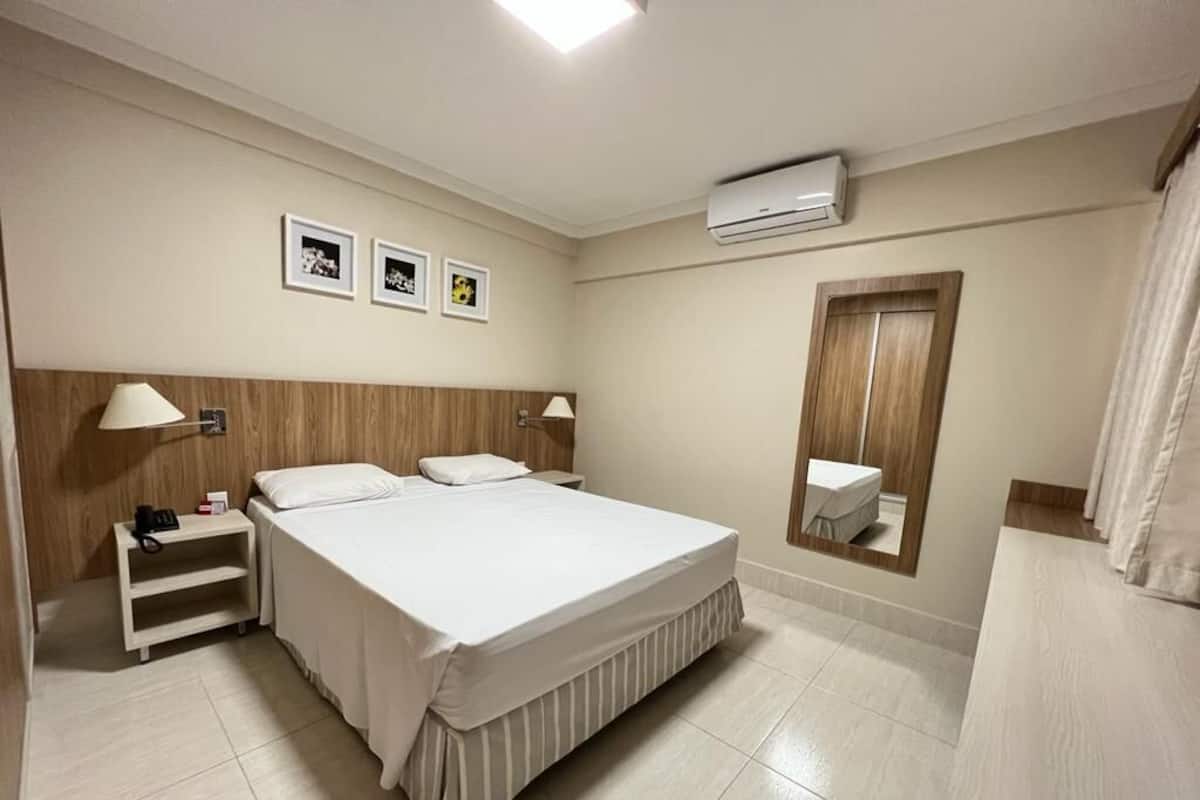 A hotel room with a large bed, bedside tables, a mirror, and a wall-mounted air conditioner.