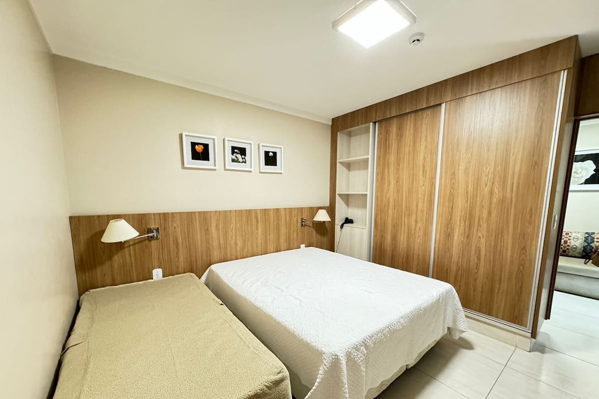 A hotel room with two beds, a wooden wardrobe, a small shelf, and three framed pictures on the wall.