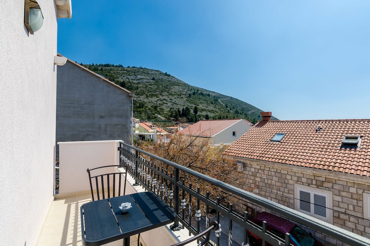 Un balcon avec une table et des chaises, donnant sur une colline et des bâtiments aux toits recouverts de tuiles rouges.