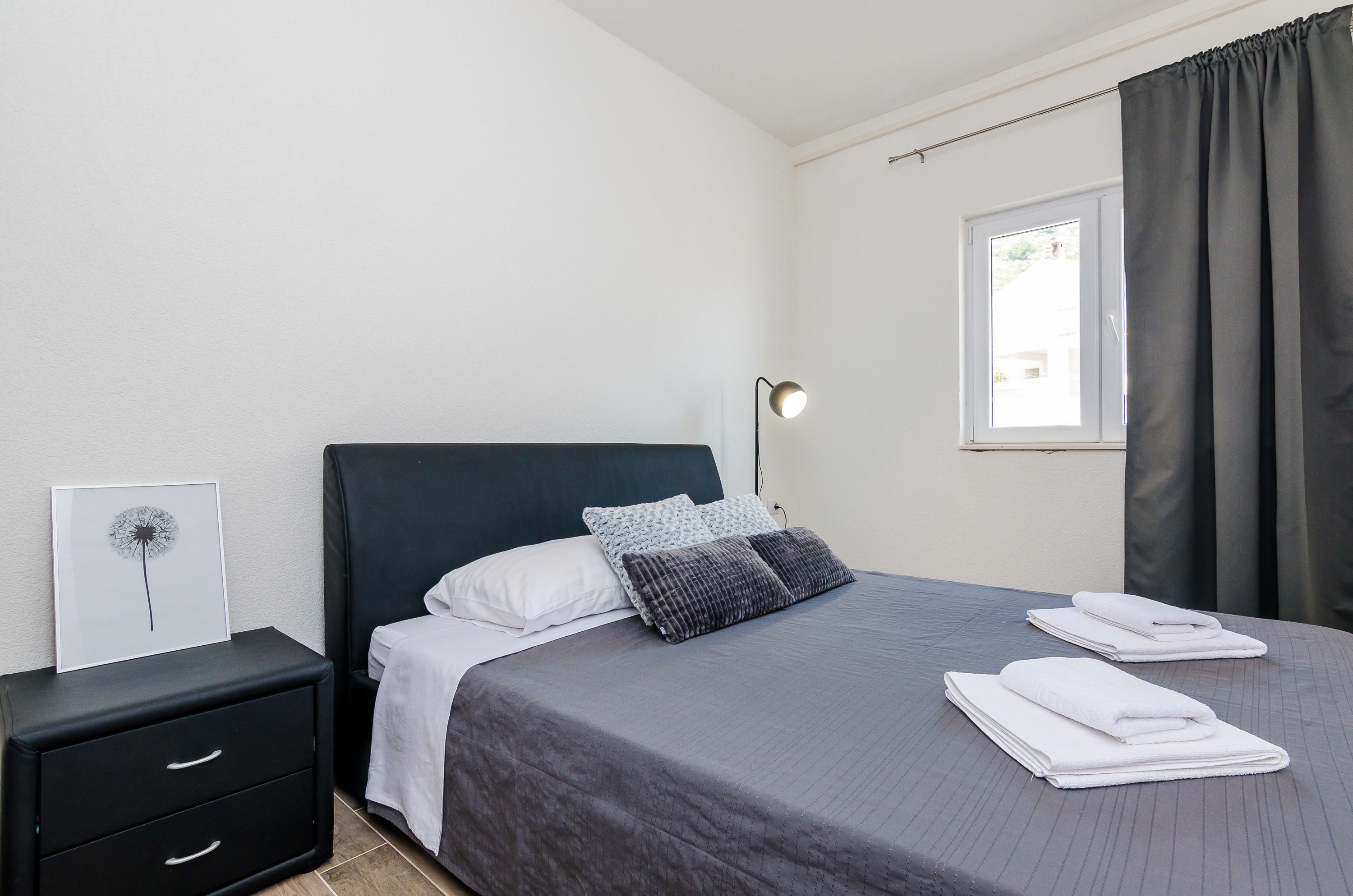 A neatly made bed with a gray bedspread and two pillows, a bedside table with a lamp, and a framed picture on the wall.
