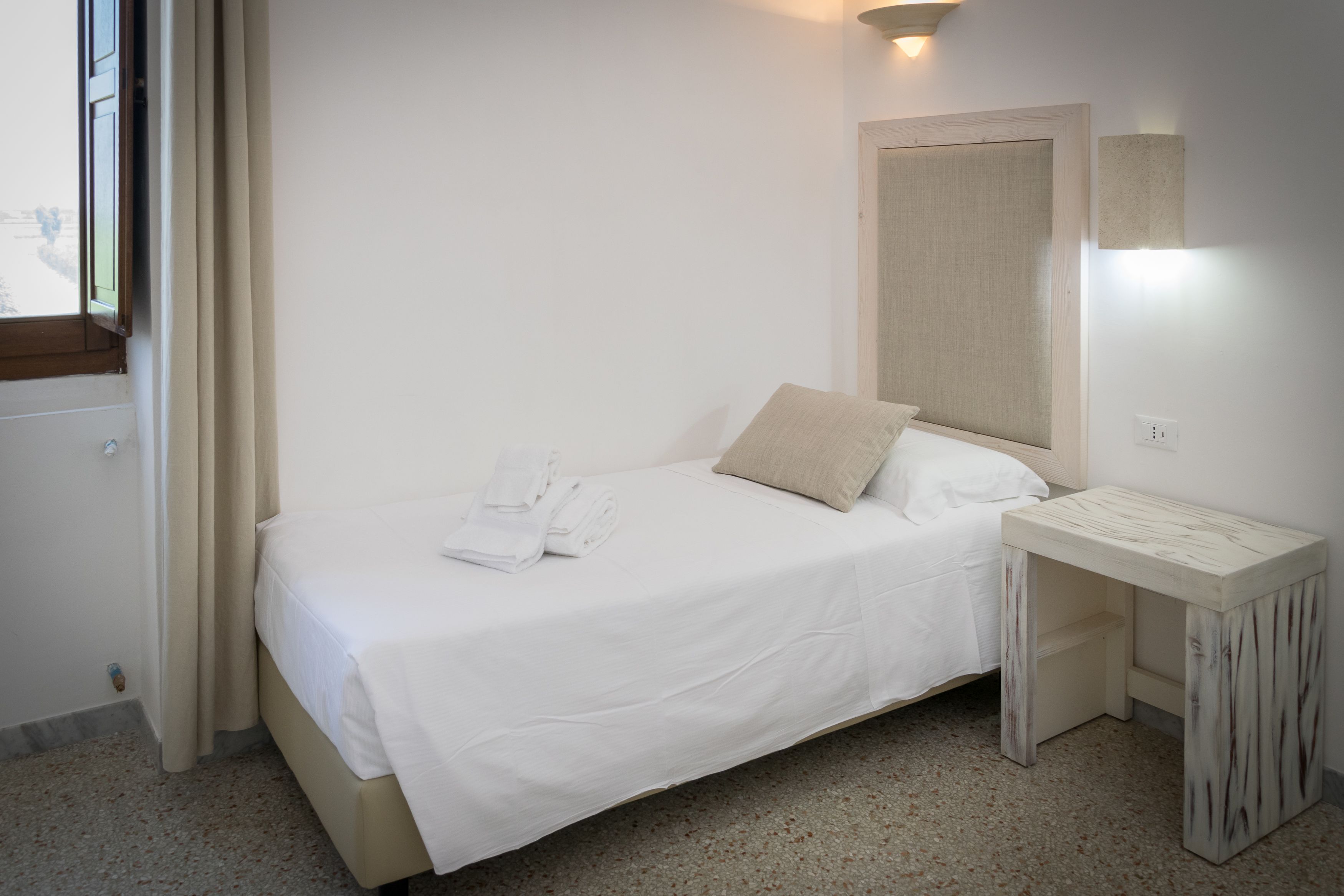 A neatly made bed with white linens, a small bedside table, and a window with curtains.