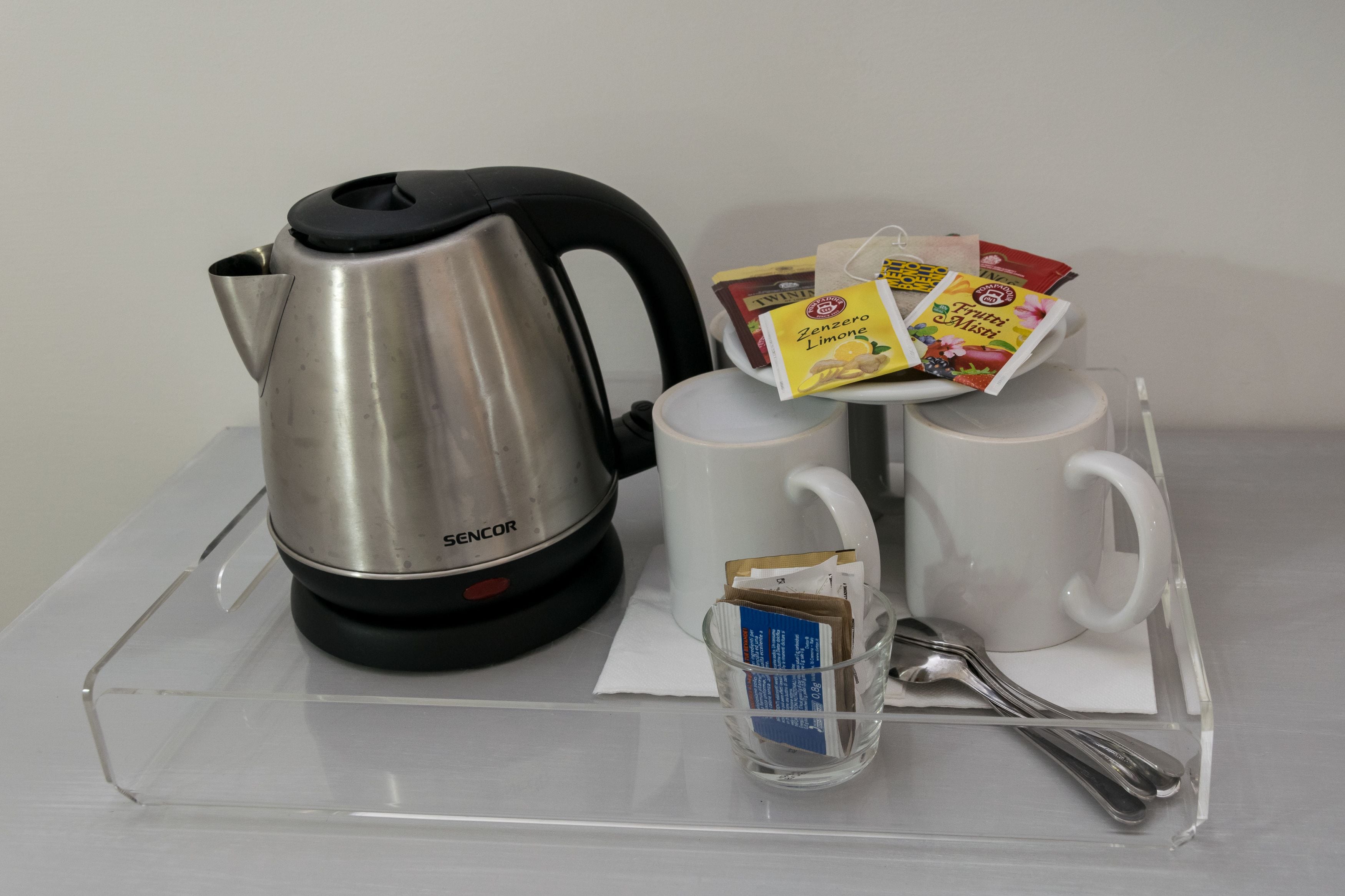A stainless steel kettle, two white mugs, a glass cup with a tea bag, and a tray with packets of tea and sugar.