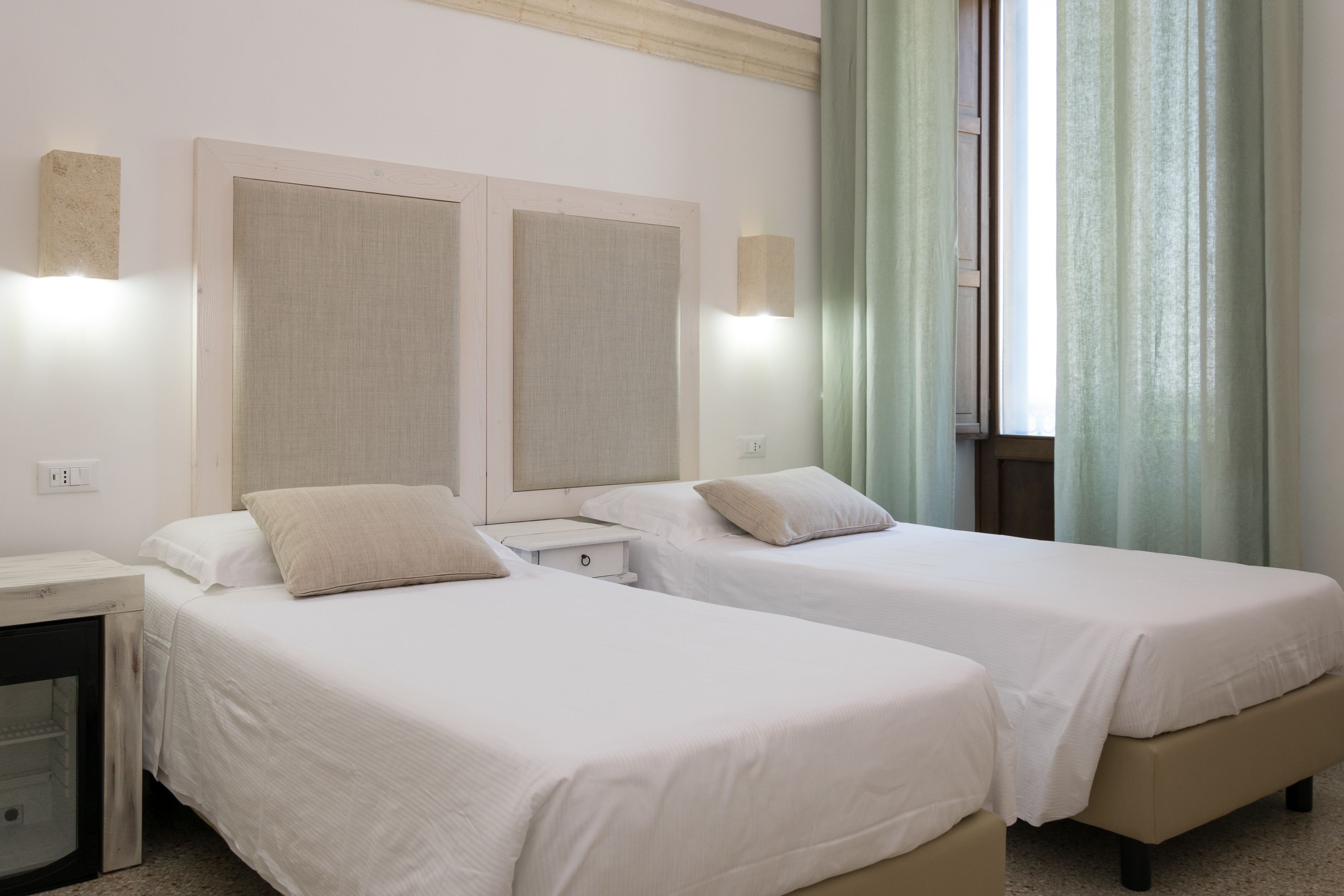 A hotel room with two single beds, a bedside table, a nightstand, and a window with curtains.