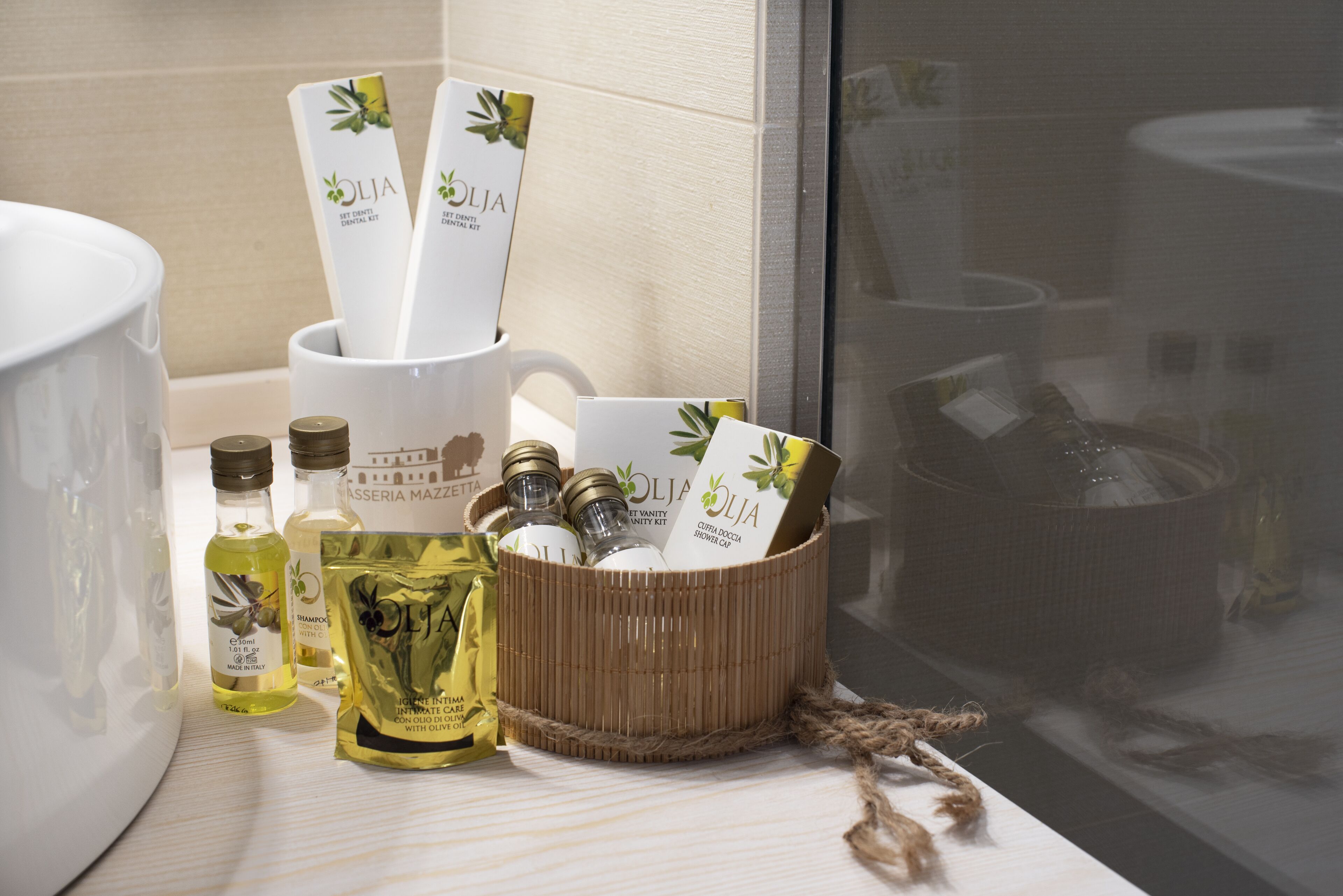 A bathroom vanity with a white sink, a large mirror, and toiletries including bottles of olive oil, a bottle of shampoo, and a bottle of lotion.