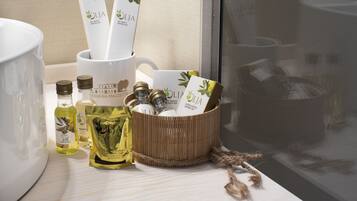 A bathroom vanity with a white sink, a large mirror, and toiletries including bottles of olive oil, a bottle of shampoo, and a bottle of lotion.