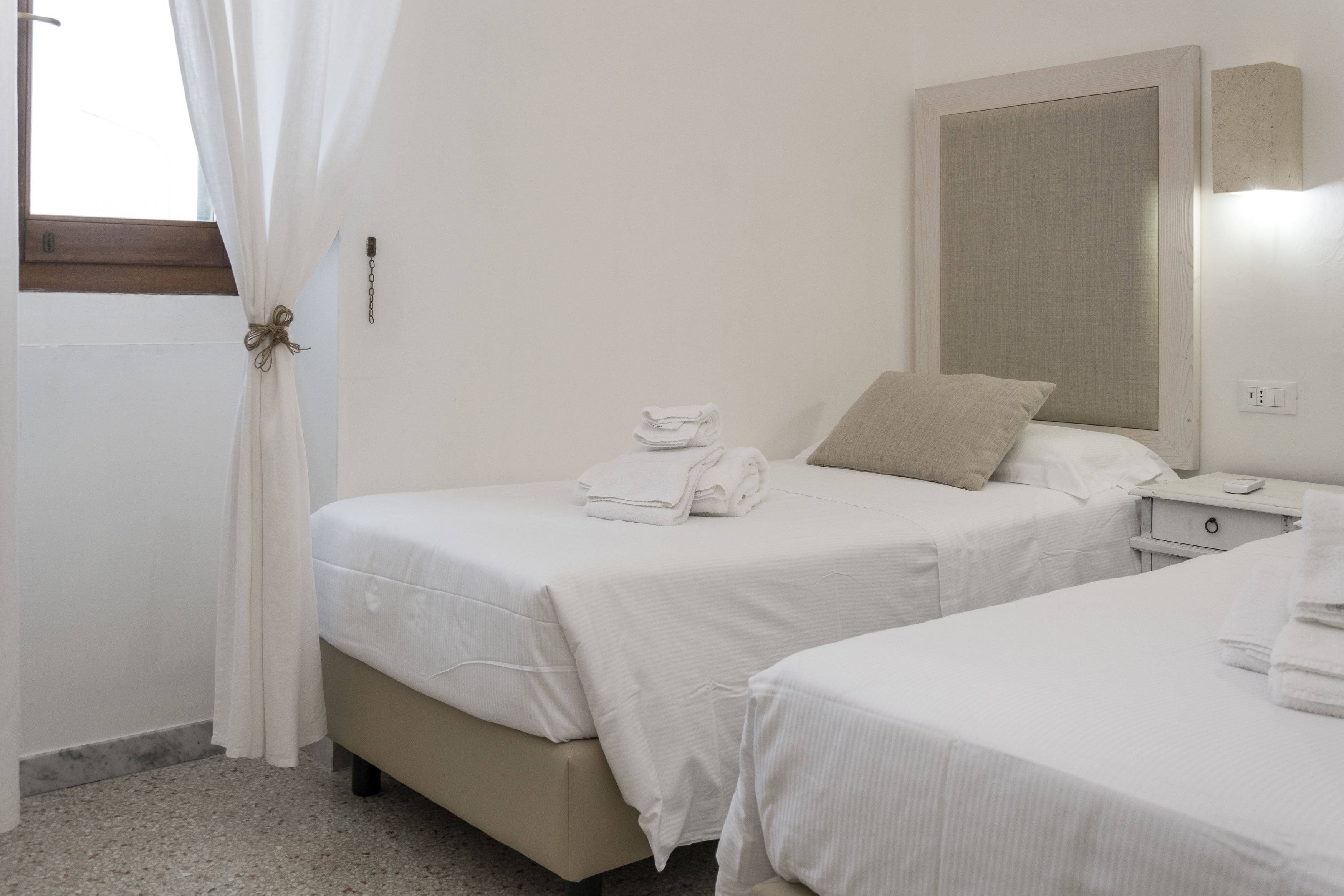 A hotel room with two beds, a bedside table, a nightstand, a window with curtains, and a wall-mounted light.