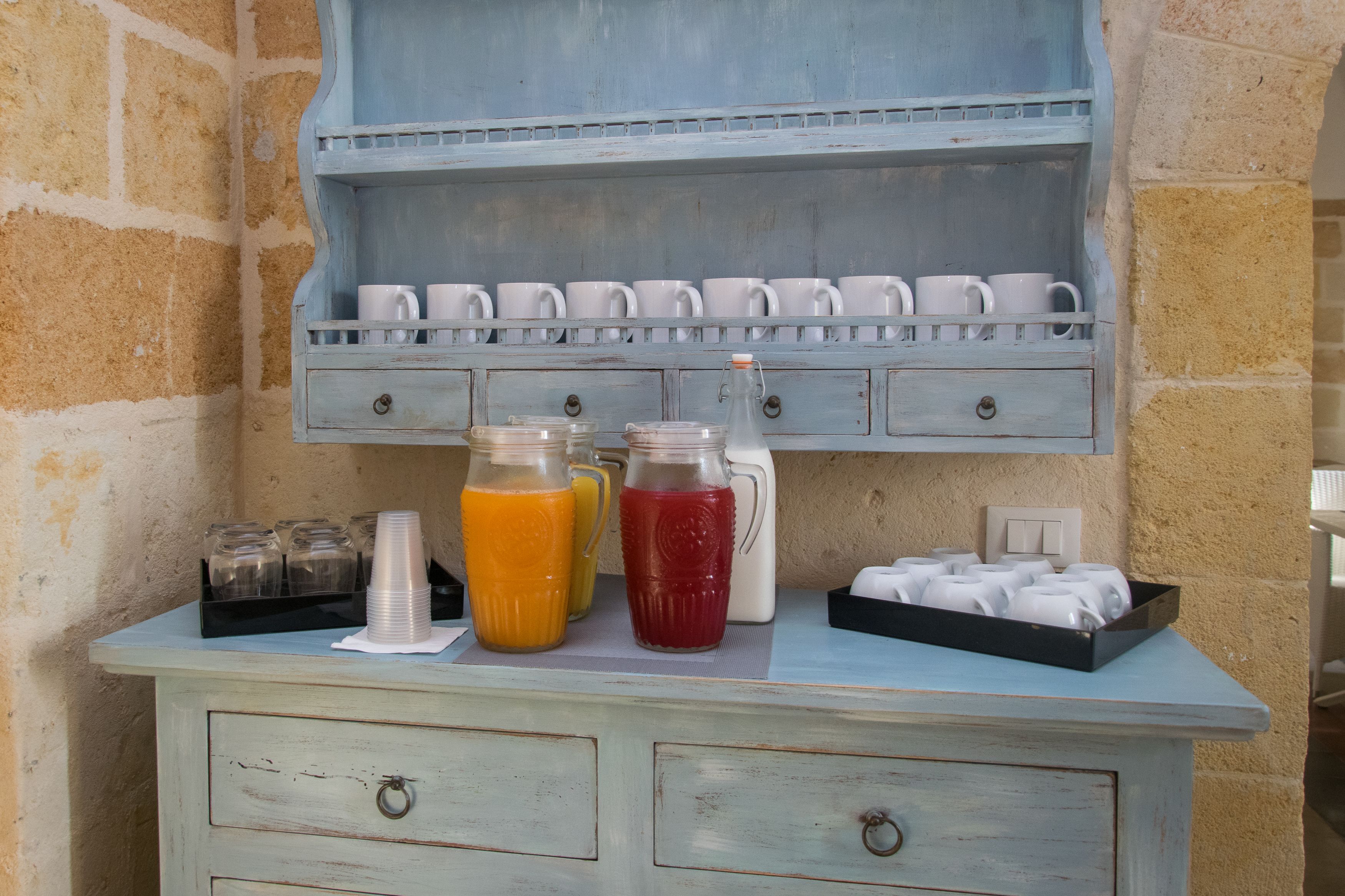A breakfast bar with a light blue color scheme, featuring a milk jug, two pitchers of orange and red beverages, and a tray of white cups.
