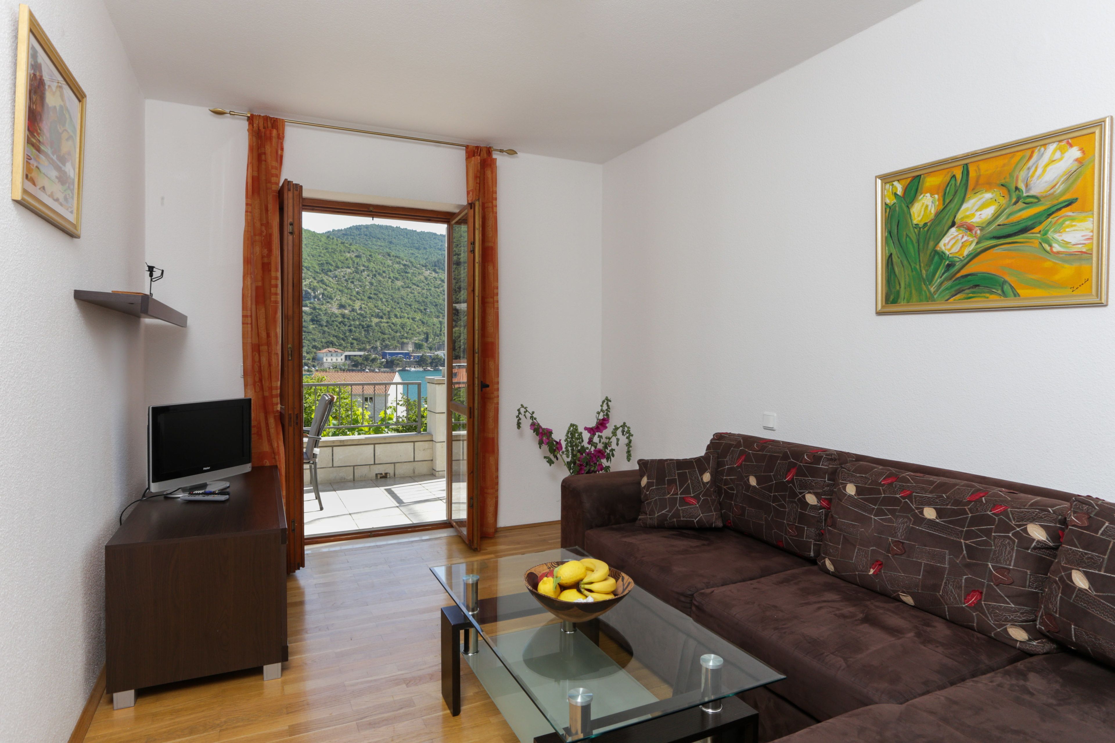 Uma sala de estar com um sofá, uma mesa de centro de vidro com frutas, uma televisão sobre um móvel e um quadro na parede.