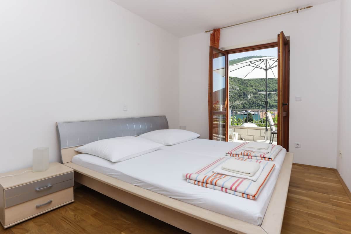 A bedroom with a bed, nightstand, and a view of a balcony with a railing and a building in the background.