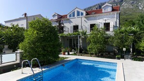 A swimming pool with a metal ladder, surrounded by lush greenery and a large building with a red-tiled roof in the background. - Villa Celenca (Dubrovnik)