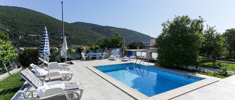 Piscina con sillones de playa y sombrillas, rodeada por un jardín ajardinado y vistas a una colina.