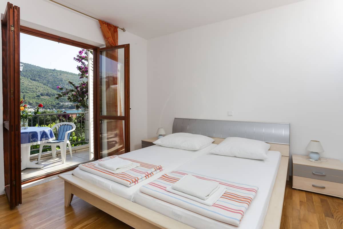 A bedroom with a large bed, a nightstand, a window with a view of a hillside, and a chair outside.