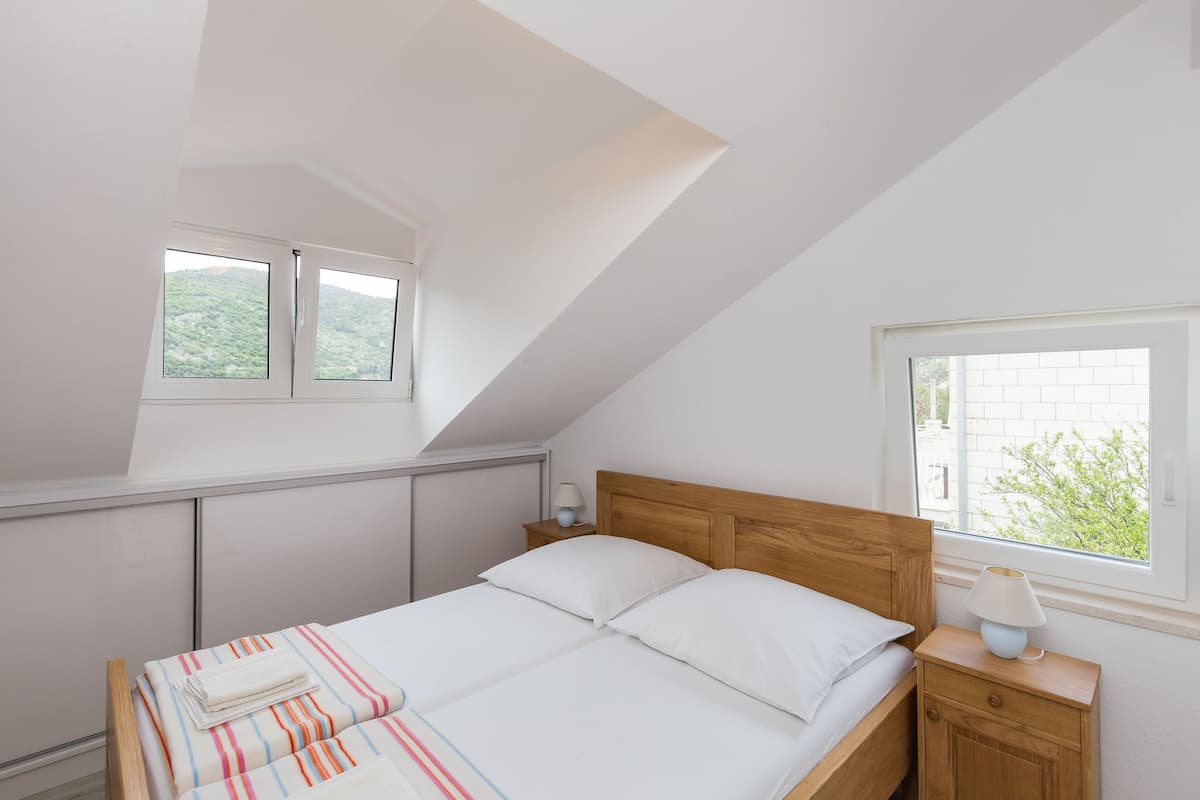A bedroom with a sloped ceiling, a bed with white bedding, a wooden headboard, a nightstand with a lamp, and a window with a view of greenery.