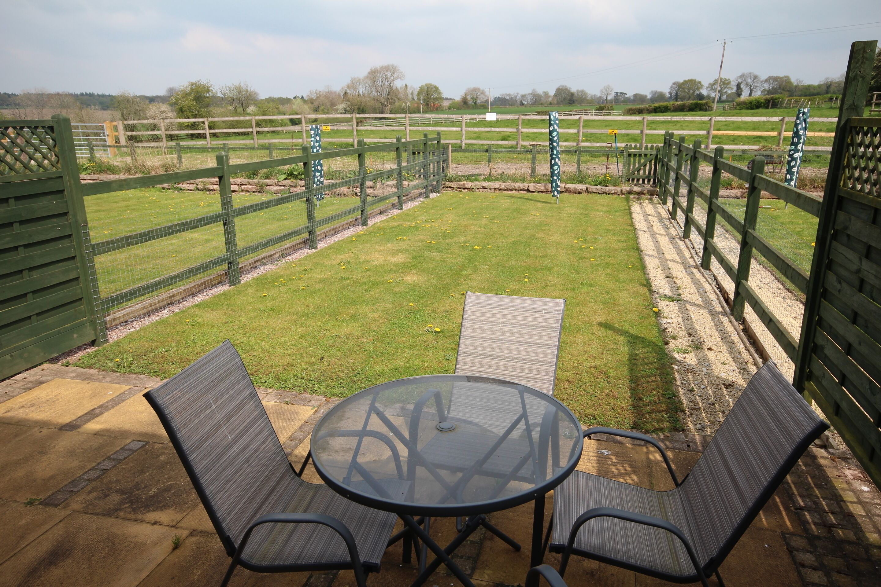 A patio with a round glass table and two chairs, surrounded by a wooden fence and a grassy field in the background.