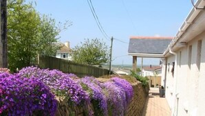 A narrow stone wall with purple flowers, a paved walkway, and a white building with a sloped roof. - Mandeley Guest House (Helston)
