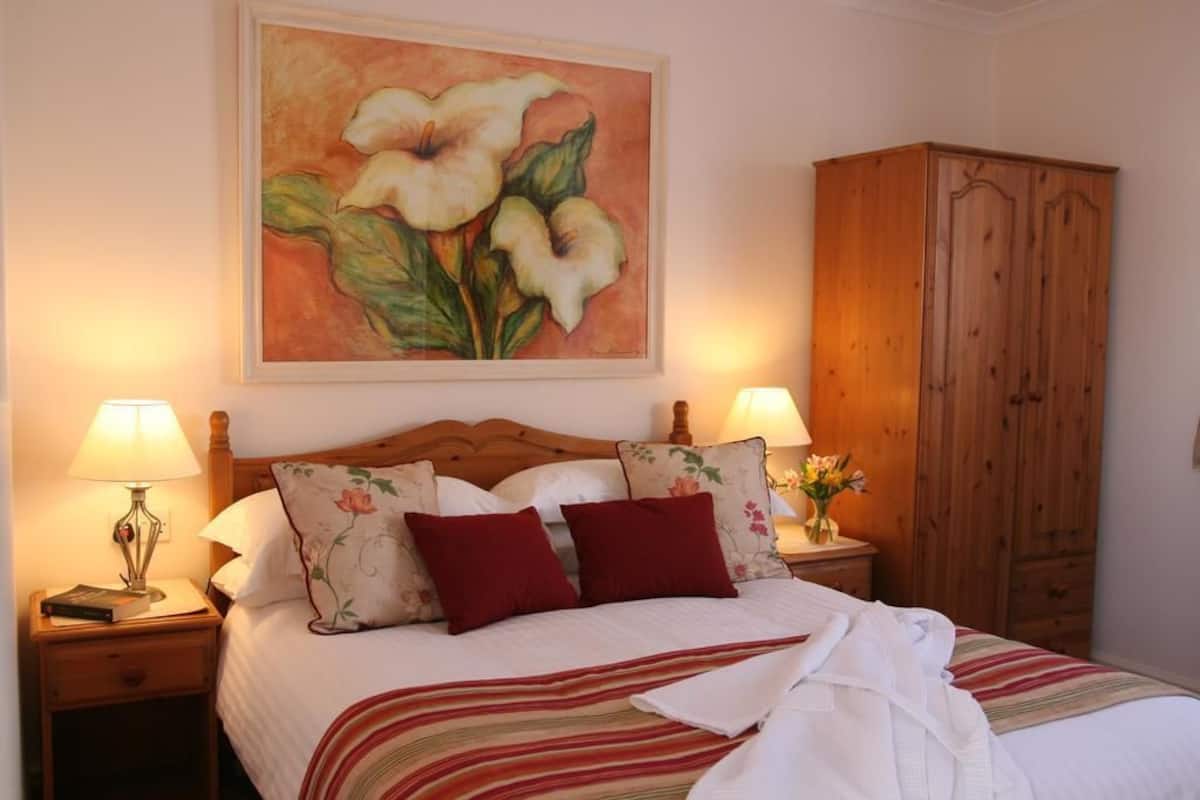 A neatly made bed with a floral pillow, a wooden wardrobe, and a painting of flowers on the wall.