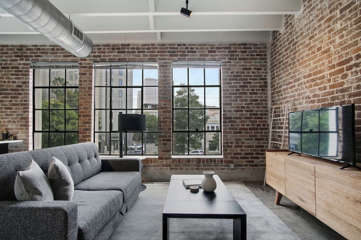 A modern living room with a grey sofa, a black coffee table, and a TV on a wooden cabinet. Large windows with a city view are visible.