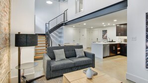 A modern living room with a grey sofa, a coffee table, and a lamp. A staircase leads to an upper level. - Roami at Motorworks (New Orleans)