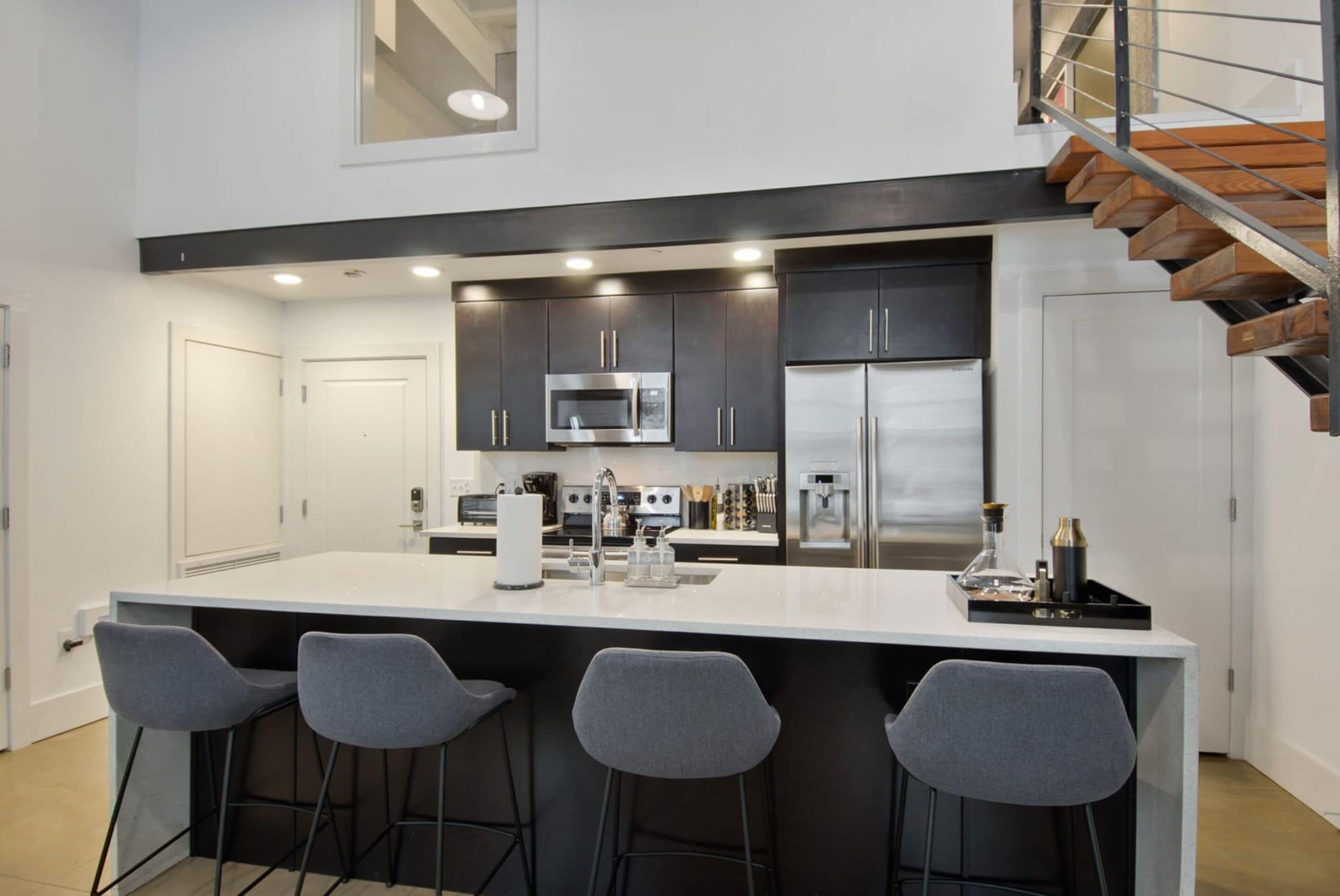 A modern kitchen with a central island, bar stools, stainless steel appliances, and a wooden staircase.