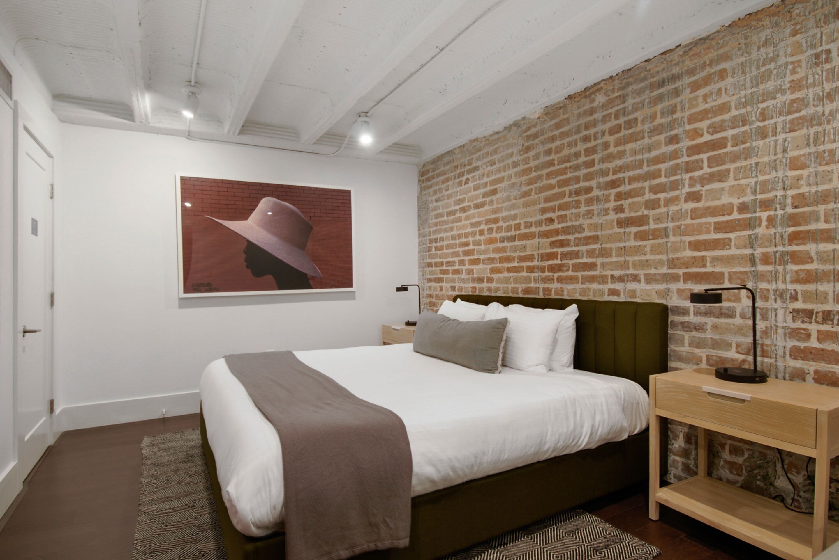 A modern bedroom with a brick wall, a large bed with a headboard, a nightstand, and a wall-mounted lamp.