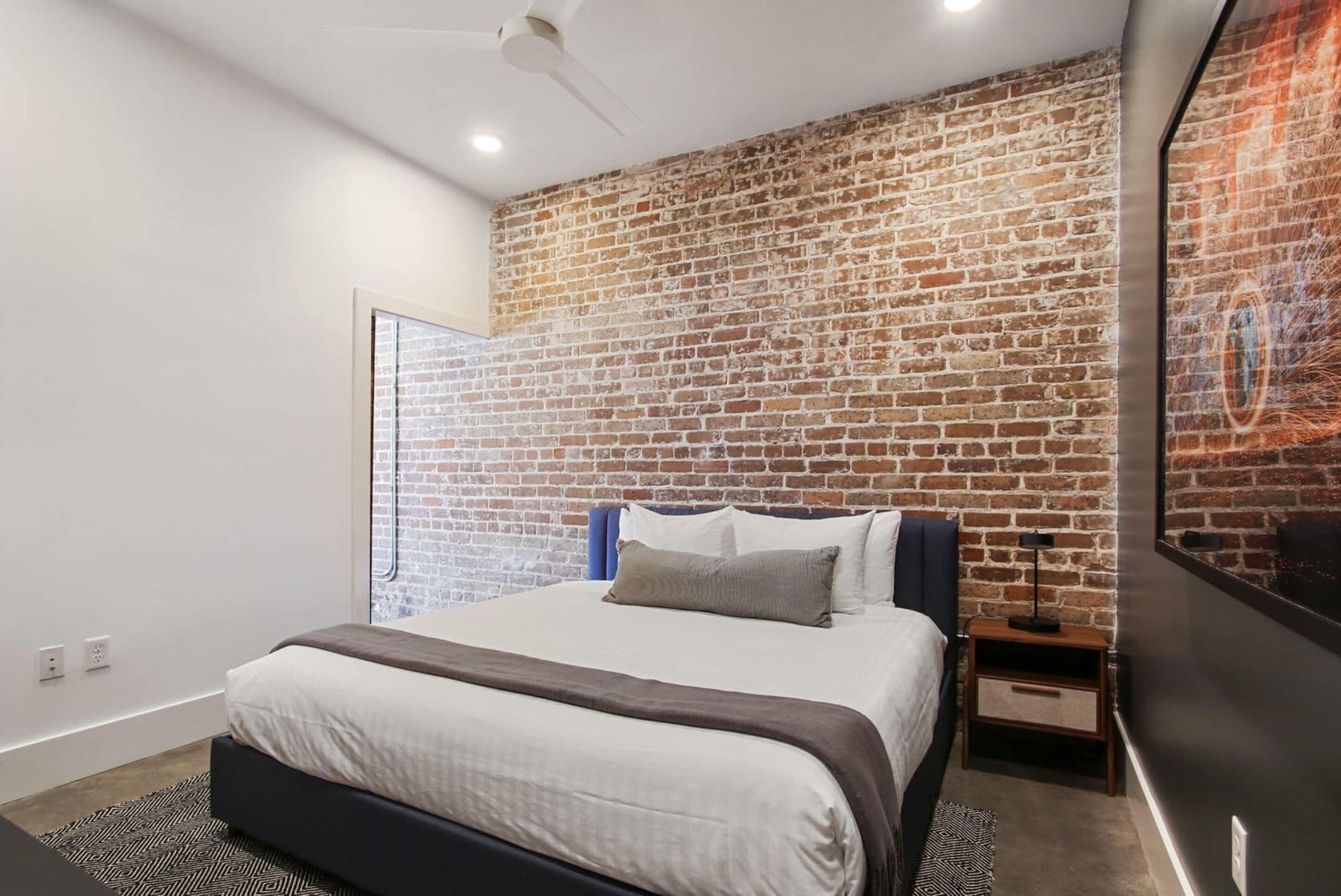 Une chambre à coucher moderne avec un mur en briques, un lit recouvert de linge de maison blanc, une table de chevet et un ventilateur de plafond.