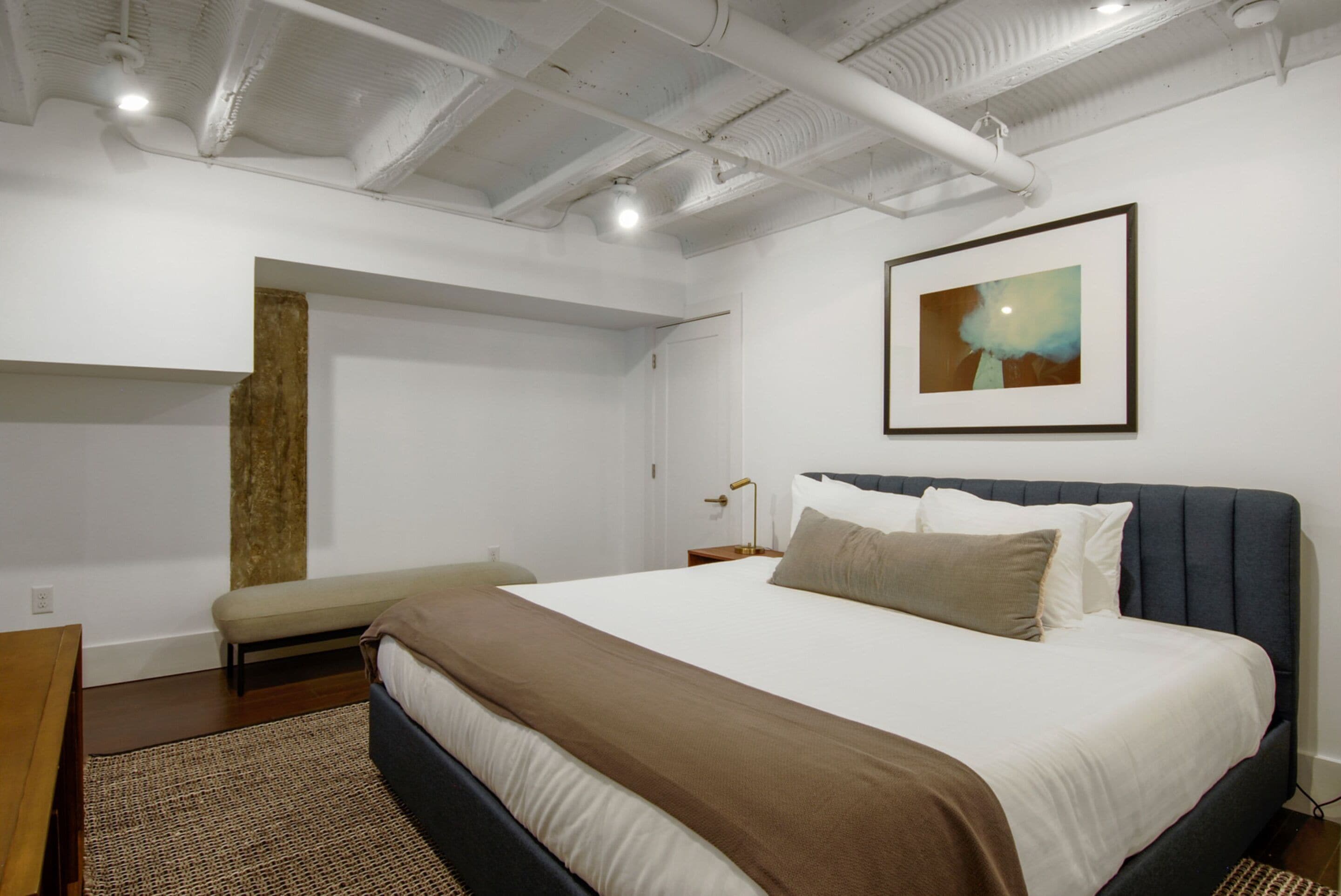 A modern bedroom with a large bed, a bench, a nightstand, and a framed picture on the wall.