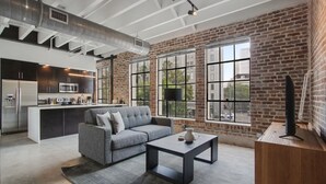 A modern kitchen with dark cabinetry and stainless steel appliances, a gray sofa with a vase, and large windows with a city view. - Roami at Motorworks (New Orleans)