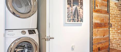 A laundry room with two white Samsung washing machines, a door with a rustic wooden panel, and a framed artwork on the wall.