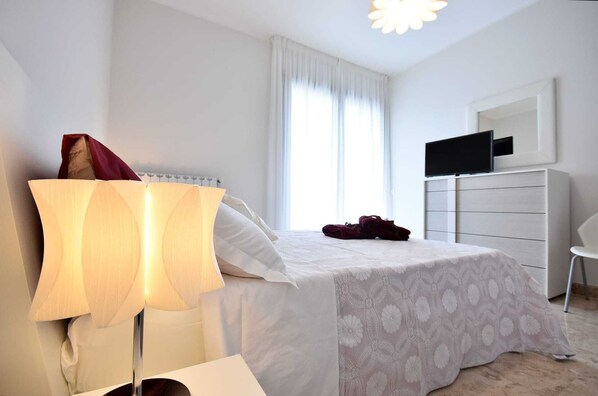 Un dormitorio con cama, mesita de noche, lámpara, televisor, cómoda y silla.