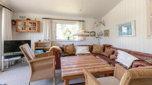 Uma sala de estar acolhedora com um sofá, cadeiras de vime, uma mesa de centro de madeira, uma televisão e uma janela com cortinas.
