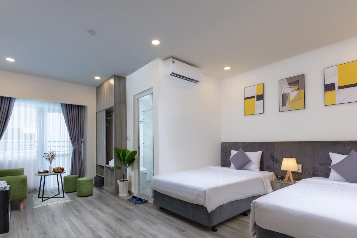 Quarto de hotel com duas camas, uma poltrona verde, uma mesinha com um vaso e uma planta em vaso.