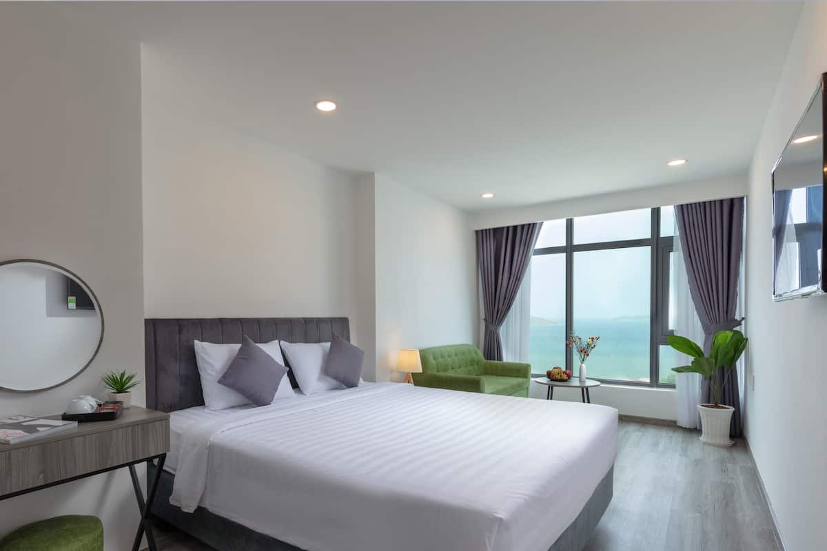 Quarto de hotel com uma cama grande, uma escrivaninha com espelho, uma cadeira verde, uma janela com vista e uma planta em vaso.
