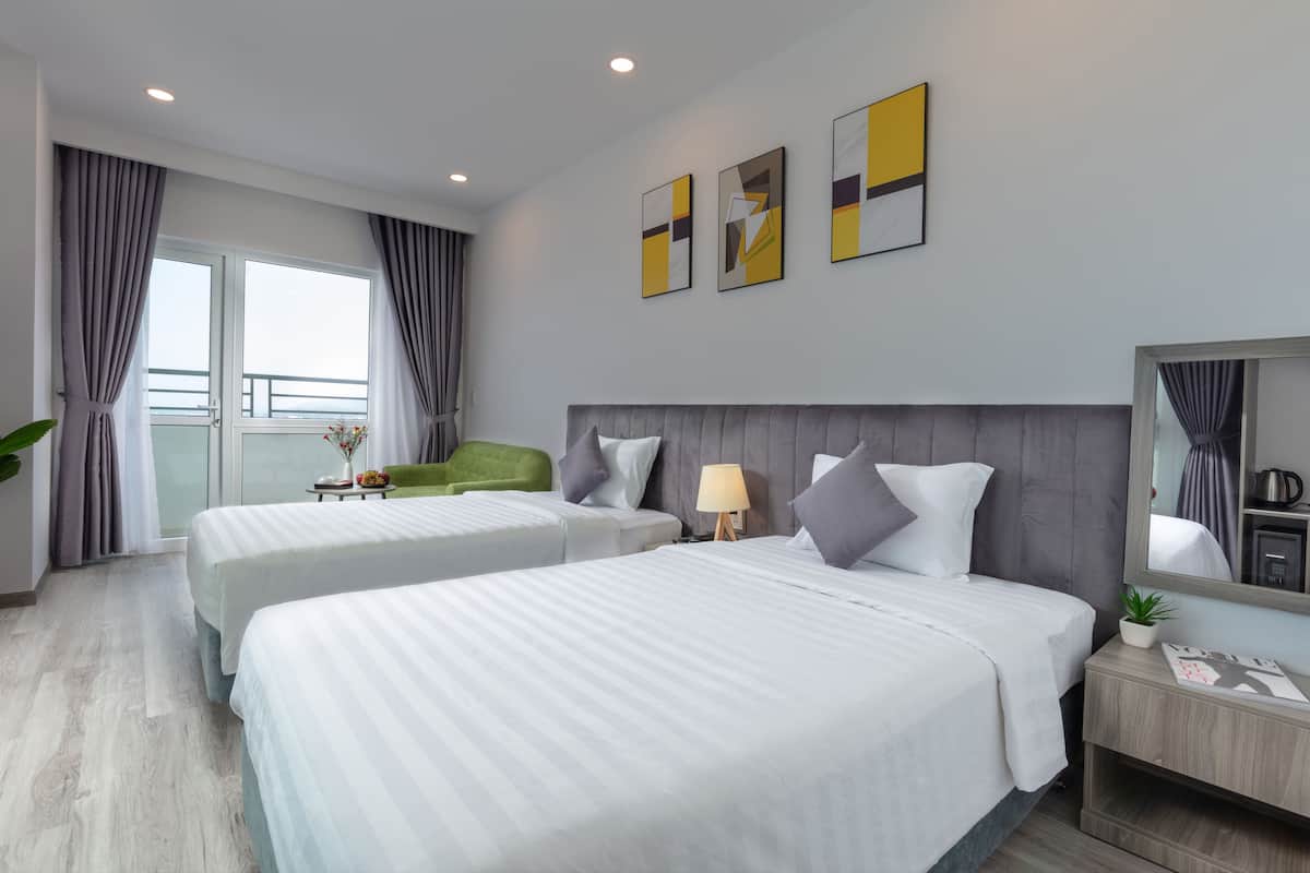 Quarto de hotel com duas camas, uma cadeira verde, uma pequena mesa com abajur e obras de arte na parede.