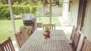 A wooden patio table with six chairs, surrounded by a garden with a barbecue grill and potted plants.