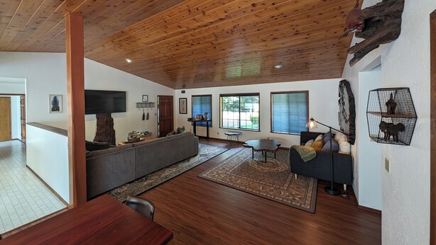 A spacious living room with a wooden ceiling, large windows, a sofa, a dining table, and a TV mounted on the wall.
